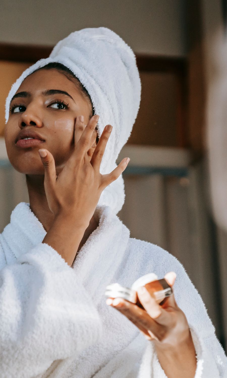 Woman in a white bathrobe and towel turban smoothing cream onto her cheek